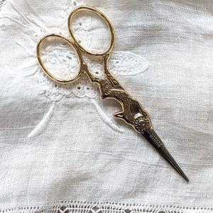 May include: Gold-colored decorative scissors with a bird-shaped handle and the word "NITHON" engraved on the blade. The scissors are lying on a white fabric with an embroidered design.