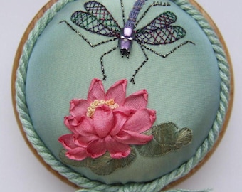 May include: A round, embroidered decorative piece featuring a dragonfly and a pink lotus flower. The dragonfly has purple and green wings, and the flower has a yellow center. The piece is set on a light blue background and framed with a green rope.