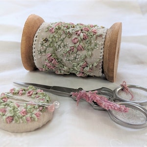 Può includere: Un rocchetto di nastro floreale rosa e verde su un rocchetto di legno. Il nastro è decorato con piccoli fiori rosa e foglie verdi. Nell'immagine sono presenti anche due spilli da cucito e un paio di forbici.