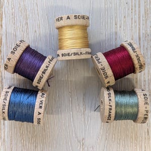May include: Five spools of silk thread in gold, purple, burgundy, blue, and green. The wooden spools are labelled "Au Ver A Soie" and "Soie/Silk-France". Suitable for embroidery and sewing projects.