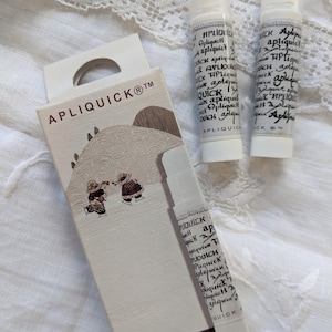 Può includere: Una scatola bianca con il testo "APLIQUICKⓇTM" e "Fabric Glue" stampato su di essa. Due tubetti bianchi di colla per tessuti con il testo "APLIQUICK" stampato su di essi sono accanto alla scatola.