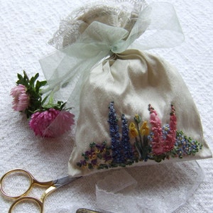 May include: A white satin pouch with a green ribbon and lace trim. The pouch is embroidered with colorful flowers, including pink, blue, and yellow.