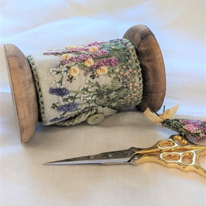 Könnte beinhalten: Eine Holzspule mit Stickgarn mit einem Blumendesign in Rosa-, Grün- und Weißtönen. Die Spule liegt auf einer weißen Oberfläche mit einer goldenen Schere.