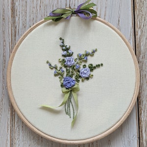 May include: A close-up of a purple and green floral embroidery on a white fabric stretched in a wooden embroidery hoop.