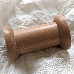 Può includere: Una bobina di legno color chiaro, con un corpo cilindrico e estremità svasate. La bobina è fatta di legno color chiaro e poggia su un tessuto bianco strutturato. Probabilmente viene utilizzata per tenere filo o lana, e misura circa 7 cm di lunghezza.