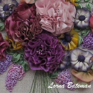 May include: A close-up of a colorful silk ribbon embroidery of a bouquet of flowers. The bouquet includes purple, pink, yellow, and white flowers. The flowers are arranged in a circular pattern and are surrounded by green leaves. The embroidery is signed "Lorna Bateman".