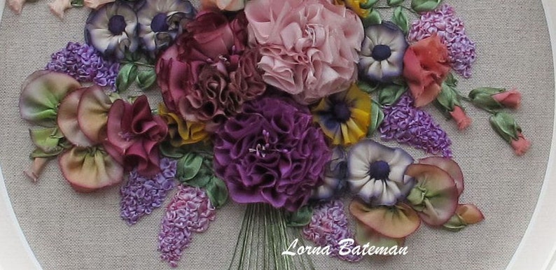 May include: A close-up of a colorful silk ribbon embroidery of a bouquet of flowers. The bouquet includes purple, pink, yellow, and white flowers. The flowers are arranged in a circular pattern and are surrounded by green leaves. The embroidery is signed "Lorna Bateman".