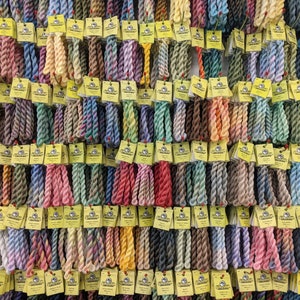 May include: A wall display of colourful embroidery floss spools, each with a yellow label featuring the brand name "DMC" and a number. The floss is arranged in rows, creating a vibrant and organised display.