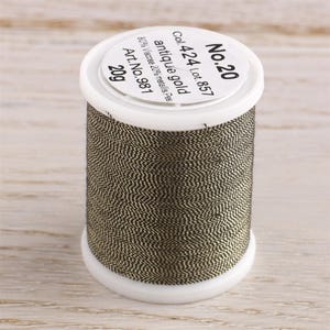K&ouml;nnte beinhalten: Eine Spule mit antikem Goldfaden, beschriftet mit "No. 20", "Col. 424 Lot. 857", "antique gold", "80% Viscose 20% metallis. Pes", "Art. No. 981" und "20g".