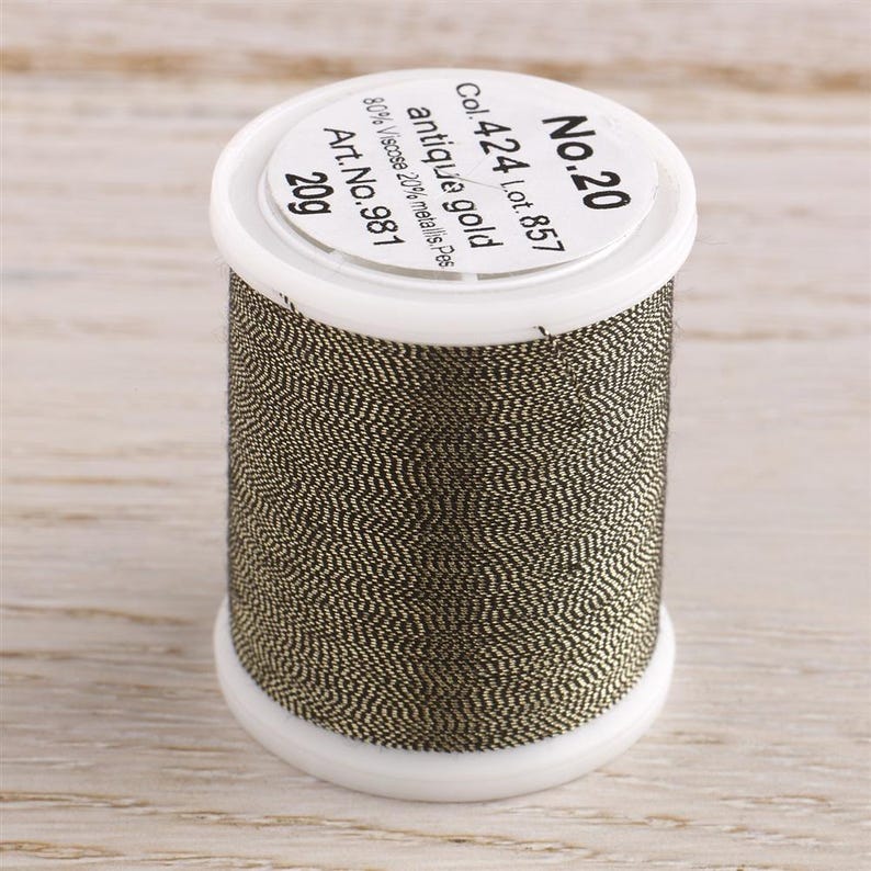 K&ouml;nnte beinhalten: Eine Spule mit antikem Goldfaden, beschriftet mit "No. 20", "Col. 424 Lot. 857", "antique gold", "80% Viscose 20% metallis. Pes", "Art. No. 981" und "20g".