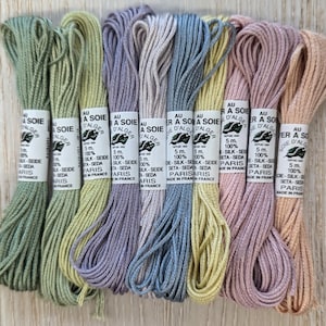 May include: Eight spools of 100% silk embroidery floss in a range of pastel shades, including green, lavender, blue, yellow, and pink. Each spool is labelled "Au fil d'Alger" and features a small illustration of a leaf. The spools are arranged in a row on a wooden surface.