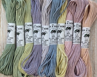 May include: Eight spools of 100% silk embroidery floss in various pastel shades, including green, lavender, blue, yellow, and pink. Each spool is labeled "Au fil d'Alger" and features a small illustration of a leaf. The spools are arranged in a row on a wooden surface.