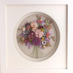 May include: A framed embroidery of a colorful bouquet of flowers in shades of pink, purple, and orange. The flowers are arranged in a circle and are surrounded by a white frame.