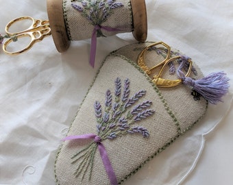 May include: A lavender-themed sewing scissor holder with a purple tassel and a pair of gold scissors. The holder is made of linen fabric and features an embroidered design of a bouquet of lavender.