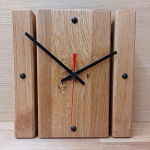 Könnte beinhalten: Eine Holzuhr mit schwarzen Zeigern und einem roten Sekundenzeiger. Die Uhr besteht aus drei Holzstücken, wobei das mittlere Stück das größte ist. Die Uhr ist an einer Holzwand montiert.