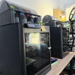 May include: Two black 3D printers with transparent front doors and top-mounted filament systems. Each unit features a digital control panel on the front. A small green alien figurine and a yellow sticky note are attached to the front of the left printer.