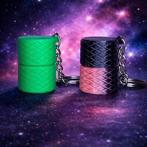 May include: Two cylindrical keychains, one green and one black and pink, with a diamond pattern. Each keychain has a silver chain and ring. The background is a colorful galaxy.