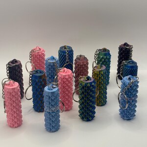 K&ouml;nnte beinhalten: Eine Sammlung zylindrischer Schl&uuml;sselanh&auml;nger mit einer texturierten, stacheligen Oberfl&auml;che. Diese Artikel sind in verschiedenen Farben erh&auml;ltlich, darunter Rosa, Blau, Gr&uuml;n und Dunkelviolett, jeweils befestigt an einem silberfarbenen Metallring.