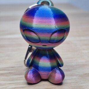 May include: A colorful alien keychain with a rainbow gradient effect. The alien figure has large, oval eyes and a small body with rounded feet. The keychain is made of a 3D-printed material and has a silver-colored clasp.
