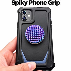 May include: A black smartphone case with a blue accent piece and a circular phone grip featuring a metallic, iridescent purple and blue spiky surface. The text Pain Stim Spiky Phone Grip is displayed above the device in bold black and purple letters.