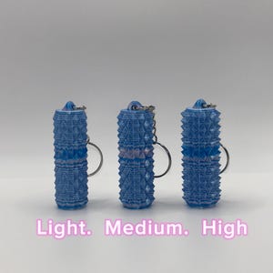 K&ouml;nnte beinhalten: Drei zylindrische, blau-wei&szlig;e Schl&uuml;sselanh&auml;nger mit einer strukturierten, geometrisch spitzen Oberfl&auml;che. Jeder Schl&uuml;sselanh&auml;nger hat oben einen Metallring. Der Text "Light. Medium. High" ist unten im Bild in Pink aufgedruckt.