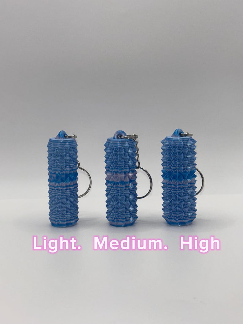 K&ouml;nnte beinhalten: Drei zylindrische, blau-wei&szlig;e Schl&uuml;sselanh&auml;nger mit einer strukturierten, geometrisch spitzen Oberfl&auml;che. Jeder Schl&uuml;sselanh&auml;nger hat oben einen Metallring. Der Text "Light. Medium. High" ist unten im Bild in Pink aufgedruckt.