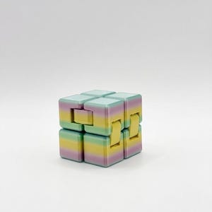 May include: A multi-jointed fidget cube toy featuring a gradient color pattern of pastel teal, yellow, and light purple horizontal stripes. The cube is constructed from several interconnected square blocks with visible hinges that allow for folding.