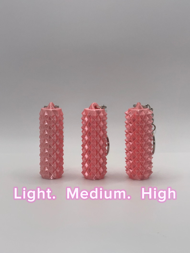 K&ouml;nnte beinhalten: Drei zylindrische, rosafarbene Schl&uuml;sselanh&auml;nger mit einer texturierten, stacheligen Oberfl&auml;che. Jeder Schl&uuml;sselanh&auml;nger hat oben einen Metallring. Der Text "Light. Medium. High" ist unten in Wei&szlig; aufgedruckt und entspricht den drei gezeigten Artikeln.