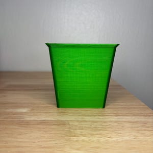 May include: A bright green, square-shaped planter with a slightly flared rim. The planter is made of a solid material and sits on a wooden surface. The background is a plain, light gray wall.