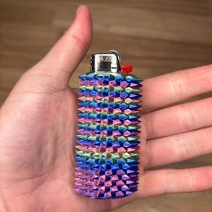 May include: A multi-colored lighter with a spiked, textured design. The colors include pink, blue, green, and purple. The lighter has a silver metal top and a red button. The lighter is held in a hand.