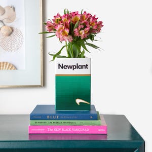 以下が含まれることがあります： 「Newplant」の黒い文字が入った、白と緑の長方形の花瓶に、ピンクと黄色の花が入っています。花瓶は、「BLUE」や「The New Black Vanguard」などの本の上に置かれています。