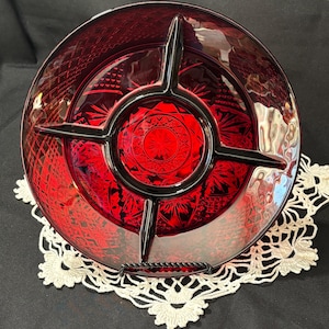 May include: A ruby red glass serving dish with a divided design. The dish has a starburst pattern in the center and a geometric pattern around the rim. The dish is supported by a black metal stand and sits on a white lace doily.