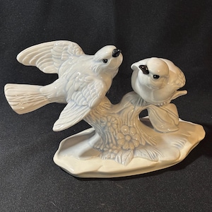 May include: A ceramic figurine of two birds perched on a stylized tree branch with flowers. The birds are light blue with black eyes and beaks. The base is a light cream color, creating a decorative accent piece.