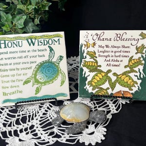 May include: Two decorative ceramic tiles with inspirational messages and sea turtle designs. One tile reads "Honu Wisdom" with a green and blue turtle. The other says "Ohana Blessing" with multiple turtles. A small turtle figurine sits in front.