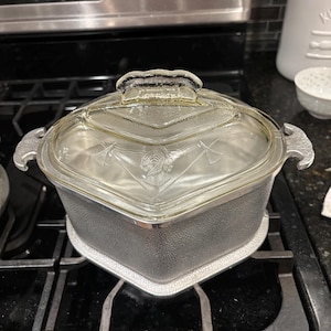 May include: A vintage, silver-colored casserole dish with a clear glass lid. The lid features a decorative handle and etched design. The dish is sitting on a black stovetop.