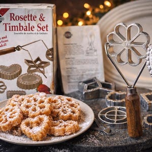May include: A rosette and timbale set with a box that reads "Rosette & Timbale Set". The set includes various metal molds, a handle, and a plate of rosette cookies dusted with powdered sugar. Strawberries are also visible.