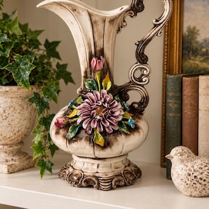 May include: A cream-colored ceramic pitcher with a dark brown antiqued finish, featuring a prominent handle and a decorative relief of purple and yellow flowers with green leaves on the front, set on a wide, ornate pedestal base.