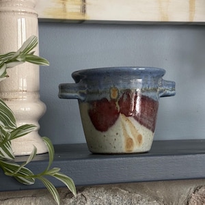 Puede incluir: Una olla de cerámica hecha a mano con borde y asas azules. La olla presenta una base crema moteada con detalles granate y bronceados. La olla se exhibe en un estante azul oscuro, mostrando su diseño rústico único.