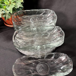 May include: Three clear glass oval dishes with a textured, wavy design. Each dish features a circular, starburst-like pattern in the center. The dishes are stacked, with a plant in the background.