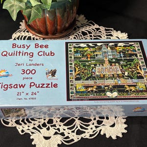 Può includere: Un puzzle da 300 pezzi del Busy Bee Quilting Club di Jeri Landers. La scatola del puzzle è azzurra e presenta una scena dettagliata di arte popolare. Il puzzle misura 53 cm x 61 cm.