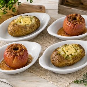May include: Four white oval ceramic dishes arranged in a square formation. Two dishes contain baked potatoes topped with a square of yellow butter and green herbs. The other two dishes contain baked apples with a brown, textured filling.