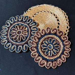May include: Three woven coasters. Two feature a floral pattern in blue, brown, and tan, while the third is a plain tan circle with a scalloped edge. The coasters are designed for placing drinks on.