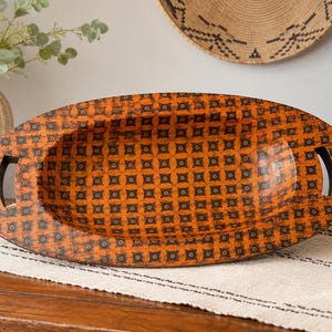 May include: An oval-shaped wooden tray featuring a vibrant orange finish with a repeating black geometric pattern of small squares and central dots. The tray includes two integrated cutout handles on the sides and a deep, concave center.