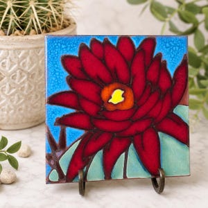 Könnte beinhalten: Eine quadratische Keramikfliese mit einer leuchtend roten Blume mit einem hellgelben und orangefarbenen Zentrum. Der Hintergrund ist ein strukturierter blauer Himmel mit einer hellgrünen Basis. Die Fliese steht auf einem dunklen Metallständer.