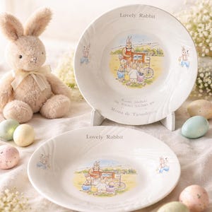 May include: A vintage-style Bunny Bowl Set with two white bowls featuring a charming rabbit illustration. The bowls have the text "Lovely Rabbit" and "Vintage Storybook Bunny Bowl Set". A plush bunny and decorative eggs complete the scene.