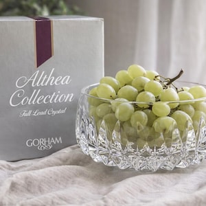 Puede incluir: Un cuenco transparente de cristal tallado lleno de uvas verdes, junto a una caja gris. La caja tiene las palabras "Althea Collection" y "Full Lead Crystal" impresas. El cuenco está sobre un paño de color claro.