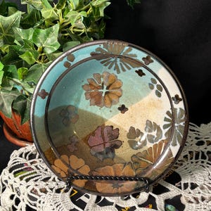 May include: A decorative ceramic plate with a floral design. The plate features a turquoise and tan color scheme with brown and purple floral patterns. The plate is displayed on a black metal stand.