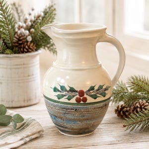 May include: A ceramic pitcher with a cream-coloured upper section featuring a hand-painted green holly leaf and red berry motif. The lower portion of the pitcher has a textured, horizontal banded finish in shades of blue, brown, and grey.