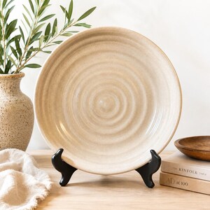 May include: A beige ceramic plate with a spiral design, displayed on a black stand. The plate is part of a home decor arrangement, with a vase of greenery and a wooden bowl in the background. The plate is approximately 12 inches in diameter.