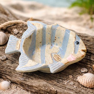 May include: A ceramic fish-shaped dish with a beige base and light blue vertical stripes. The dish has a small, round eye and a pink accent on the edge. It is resting on a piece of driftwood, with seashells and sand in the background.
